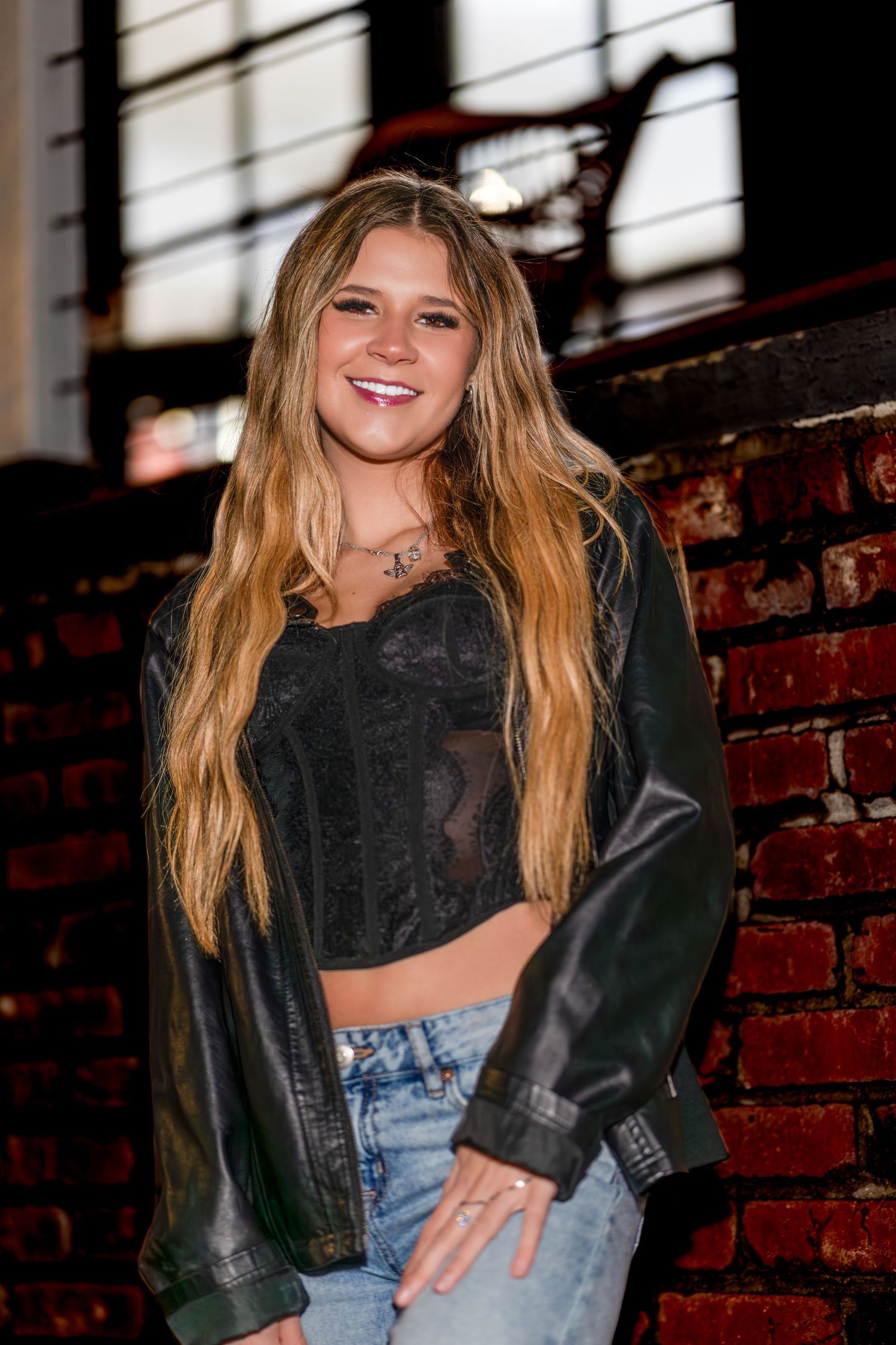 A person wearing a black crop top and denim jeans poses against a brick wall with a bright smile.