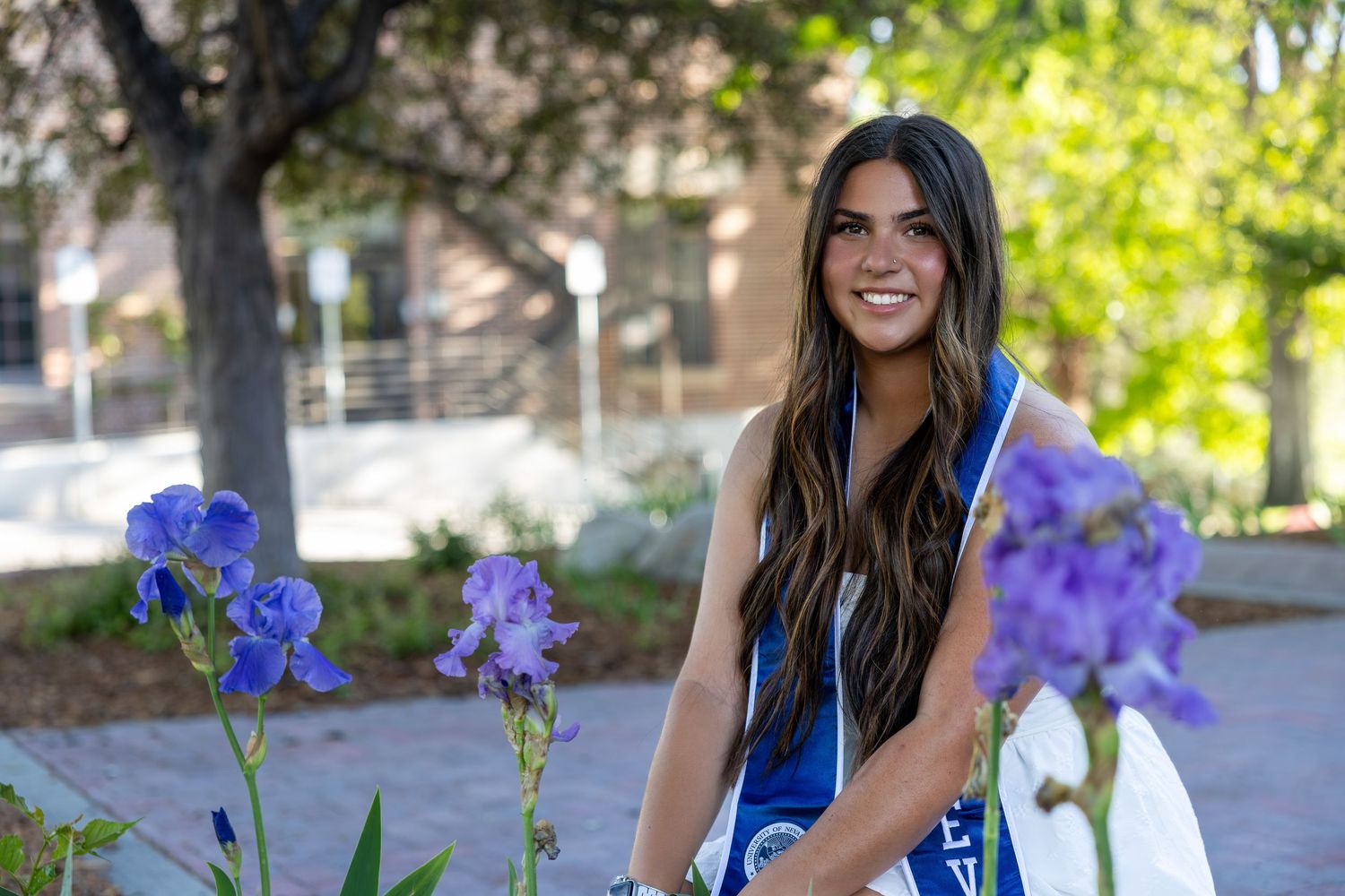 Smiling college graduate wearing a blue sash poses outdoors near purple iris flowers on a sunny campus day.