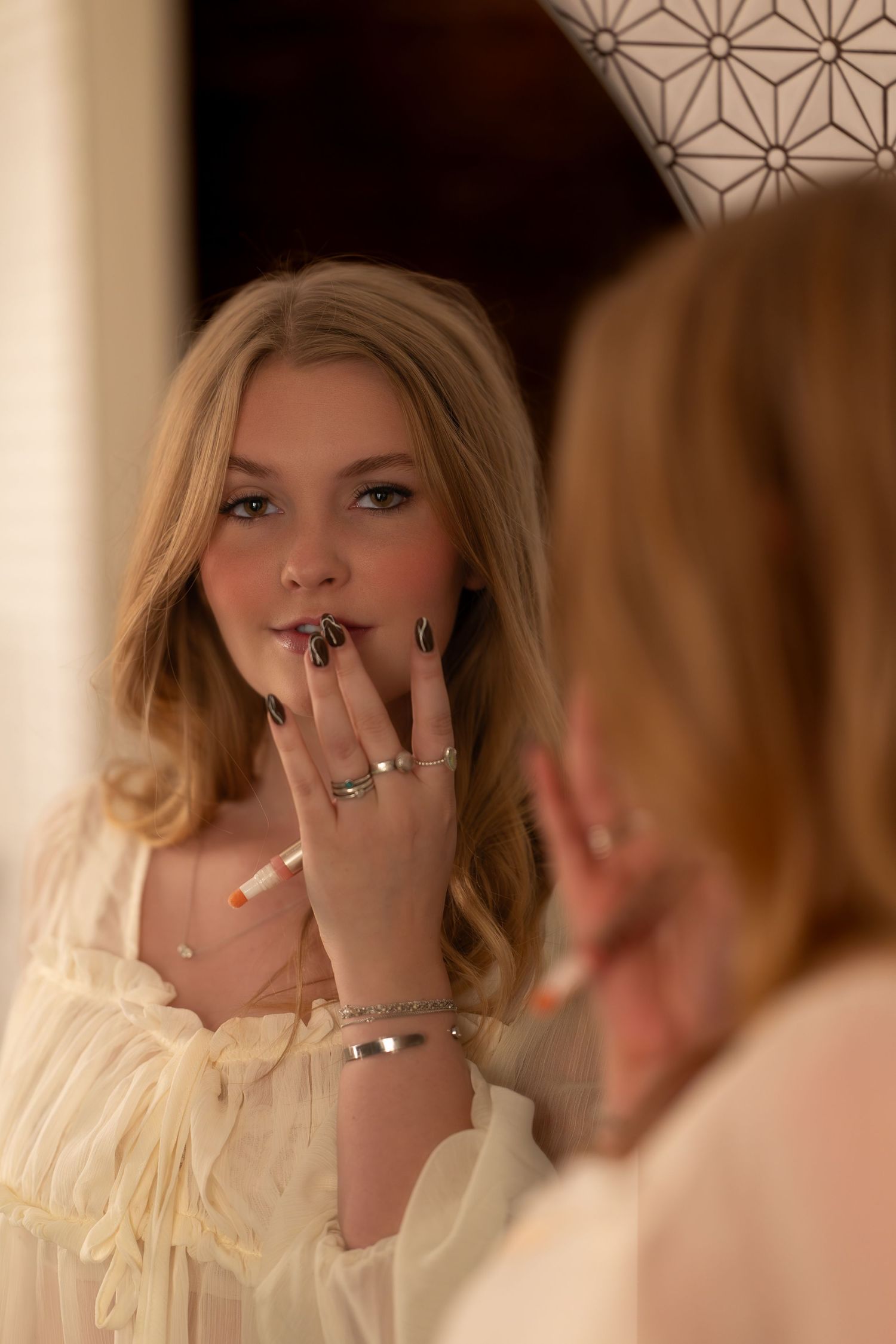 A series of mirror reflections show a person in a cream-colored outfit applying lipstick in warm lighting.