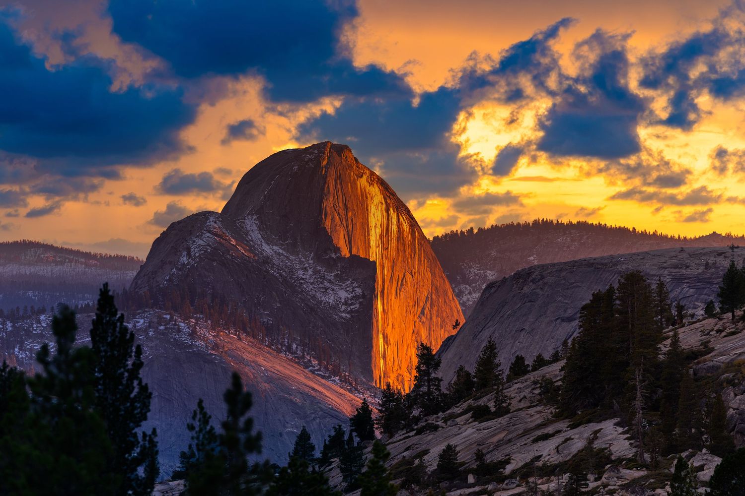 Half Dome rock formation in Yosemite National Park glows orange during a dramatic sunset with clouds in the sky.