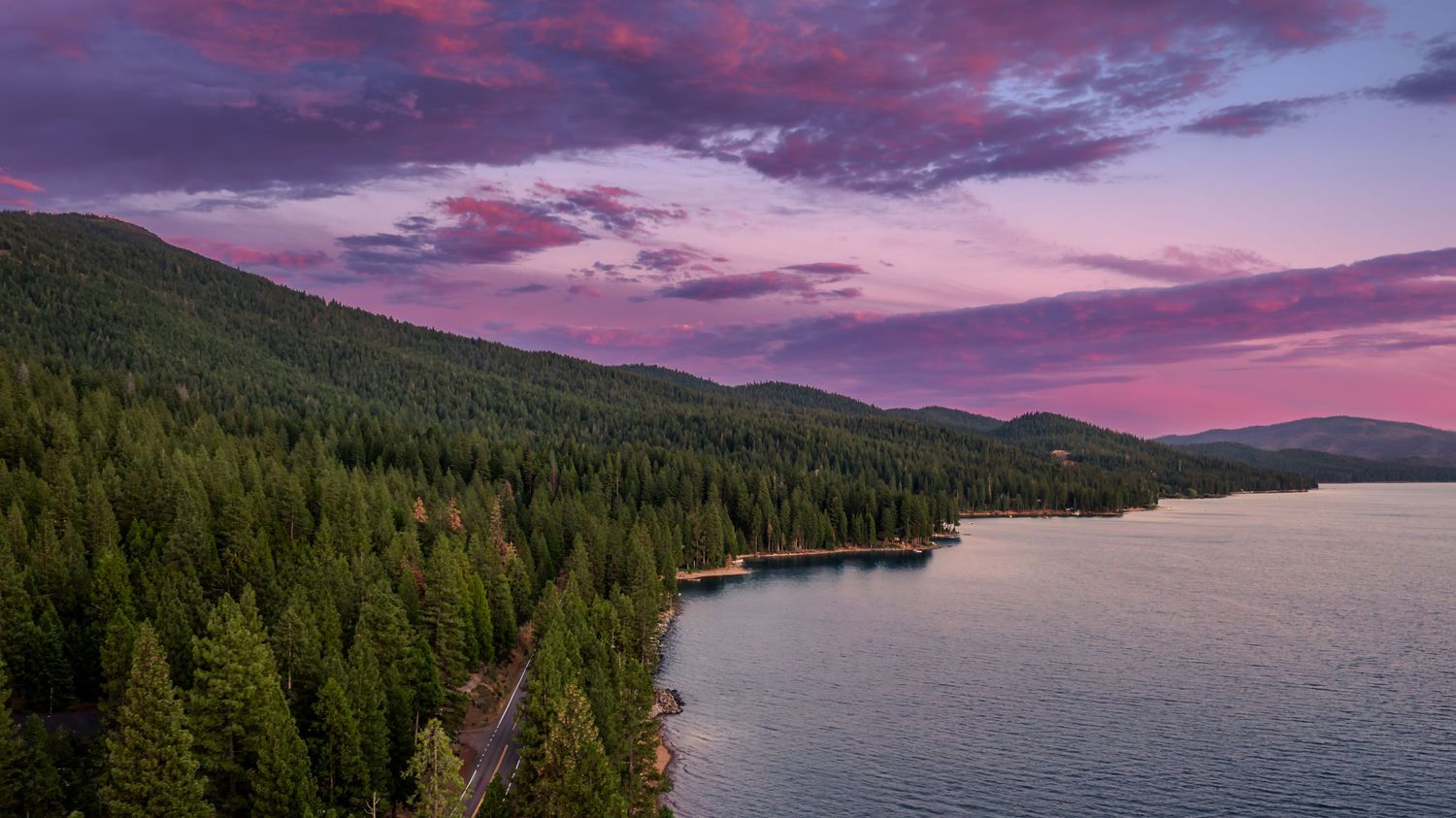 A stunning panoramic view of mountain lakes and forests under a dramatic purple and pink sunset sky.