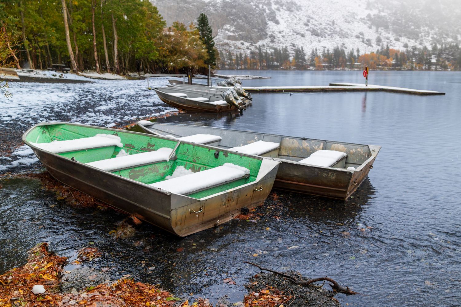 Several wooden rowboats tied up along a lake shore with autumn trees and mountains in the background.