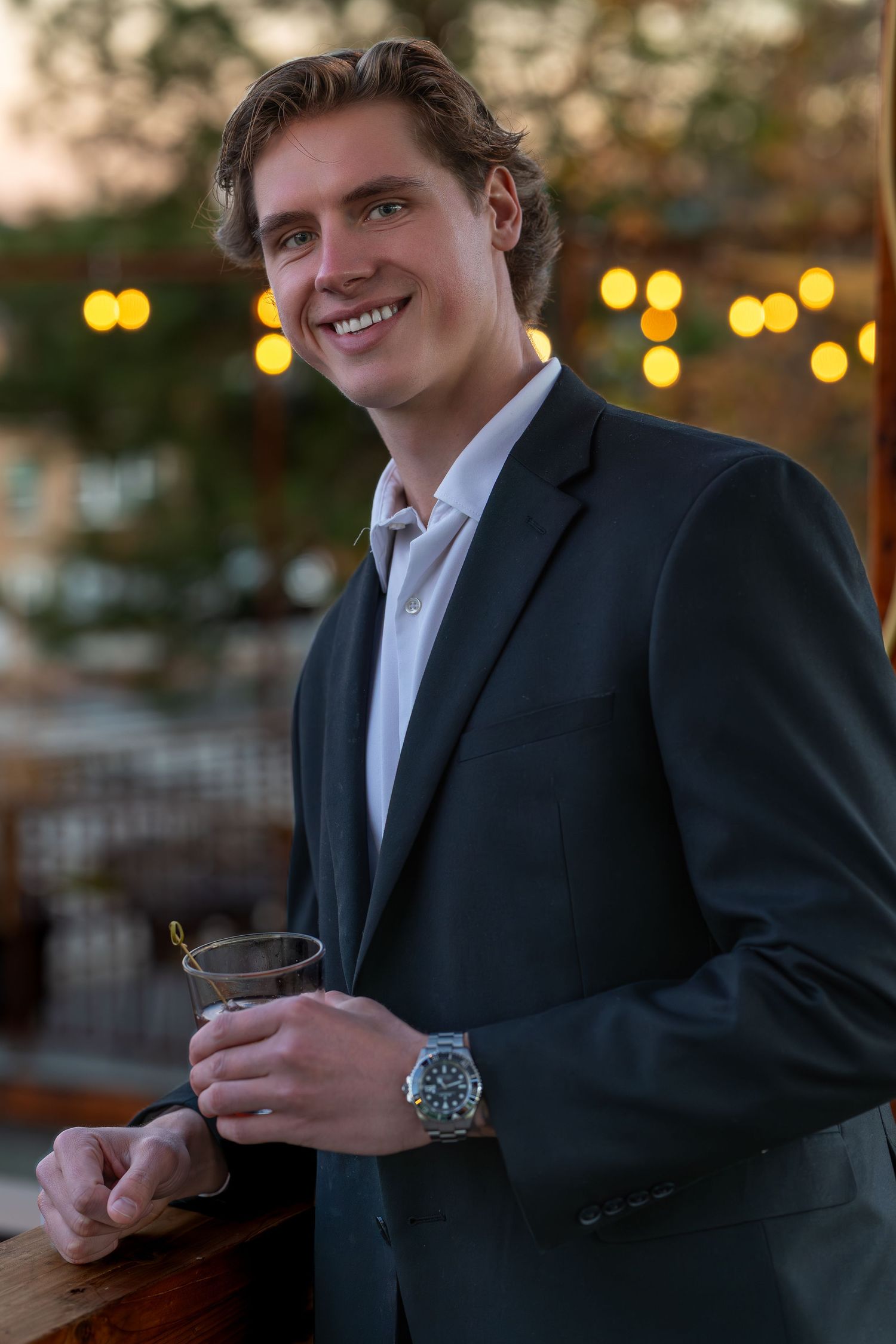Person in black suit jacket and white shirt holding drink at evening event with bokeh lights in background.
