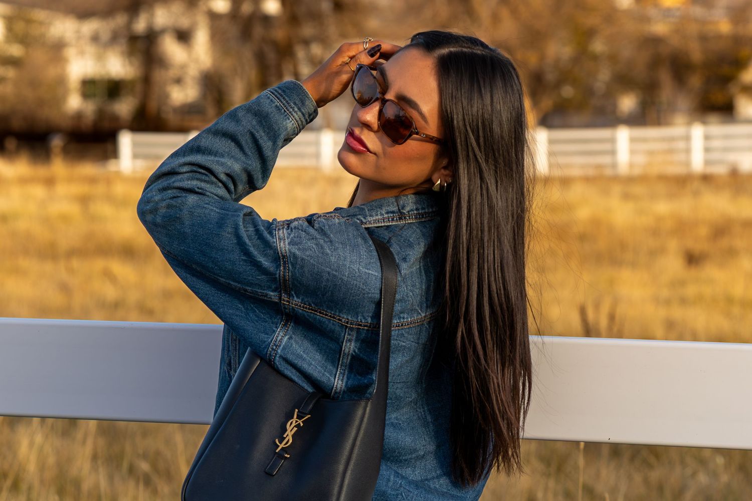 Woman in denim jacket and sunglasses posing outdoors near white fence with YSL bag.