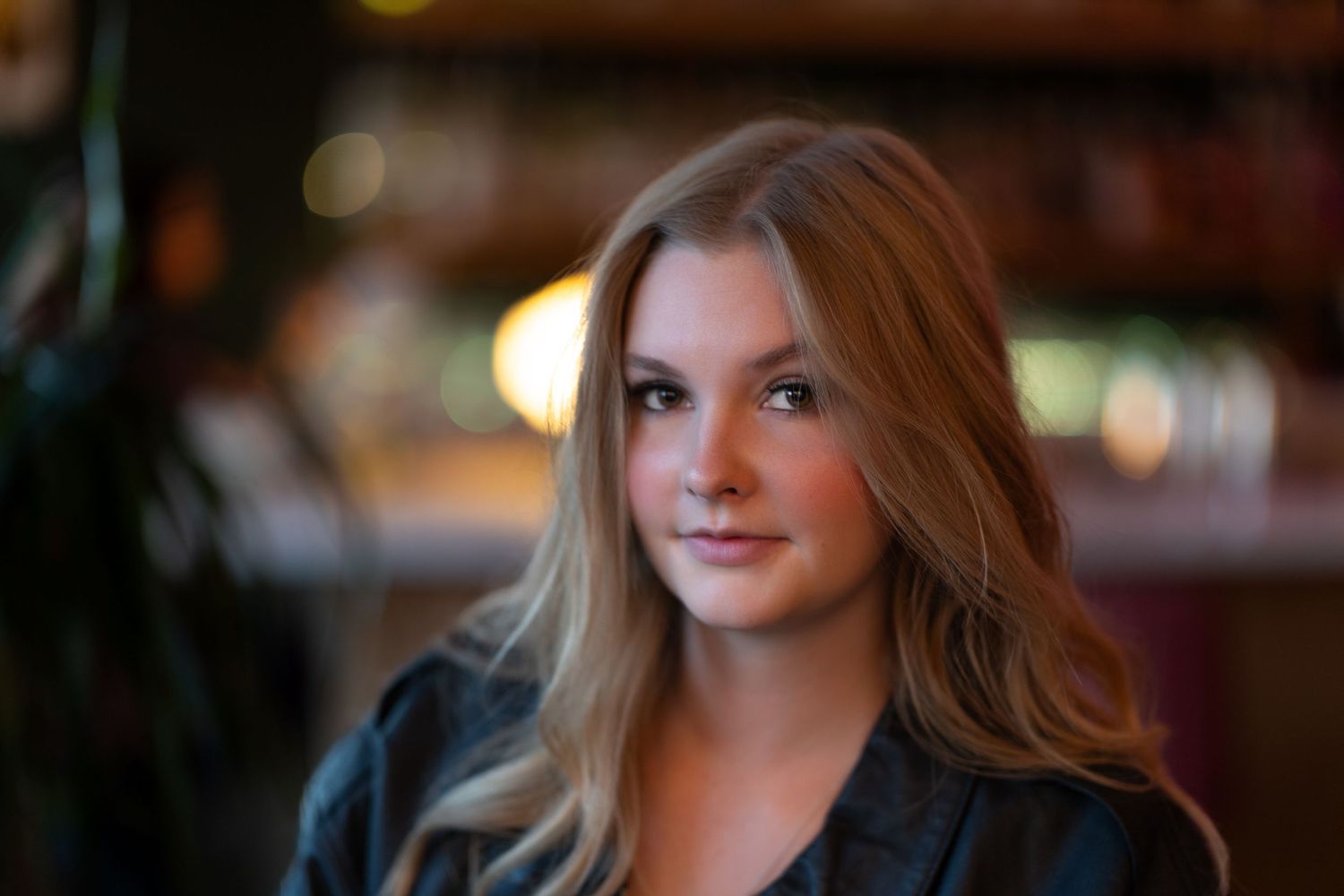 Young woman with long blonde hair posing indoors with a blurred warm-lit background.