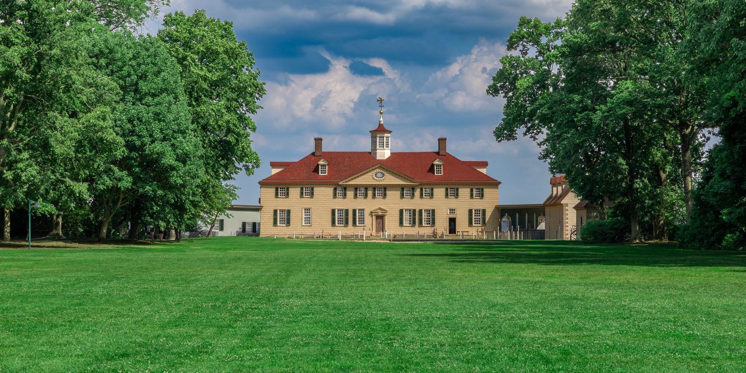 Mount Vernon estate with lush green lawn, colonial mansion, red roof, and tall trees under dramatic cloudy sky.