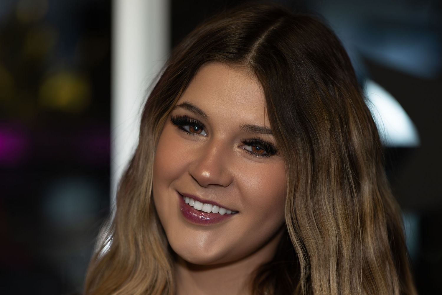 A smiling person with long ombre hair and dark eye makeup poses for a close-up portrait.