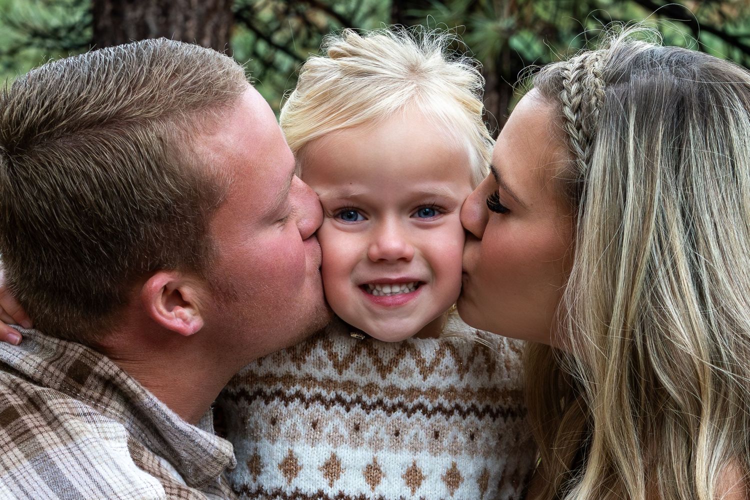 A young child smiles while receiving kisses on both cheeks while wearing a patterned sweater.