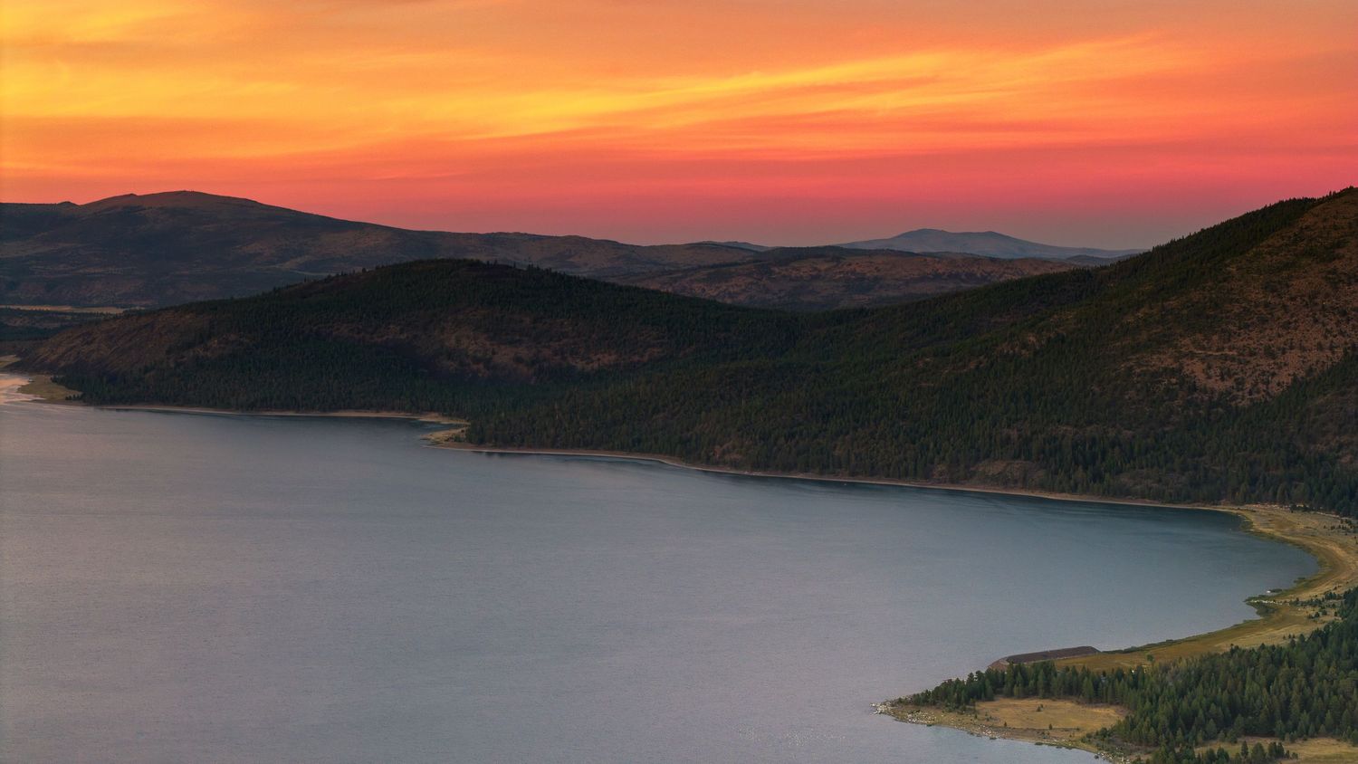Mountain lake landscape at sunset with evergreen forest and pink-orange sky.