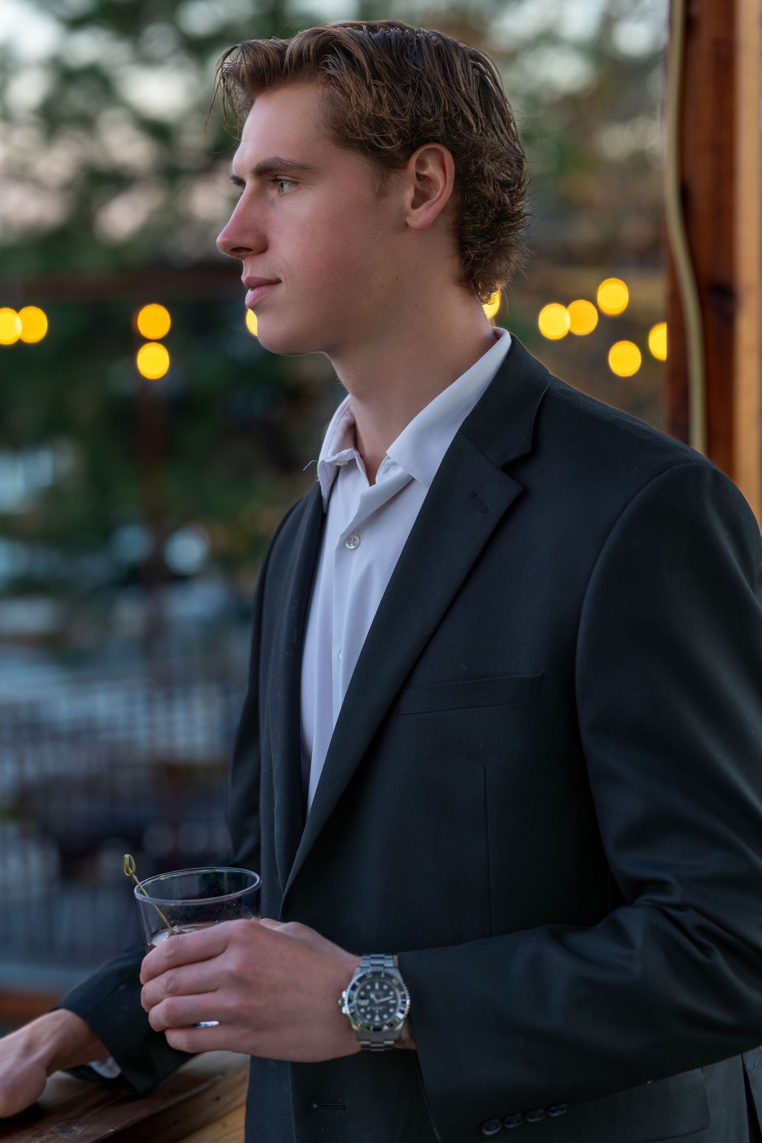 Young man in dark blazer holding a drink glass at an outdoor evening event with string lights.