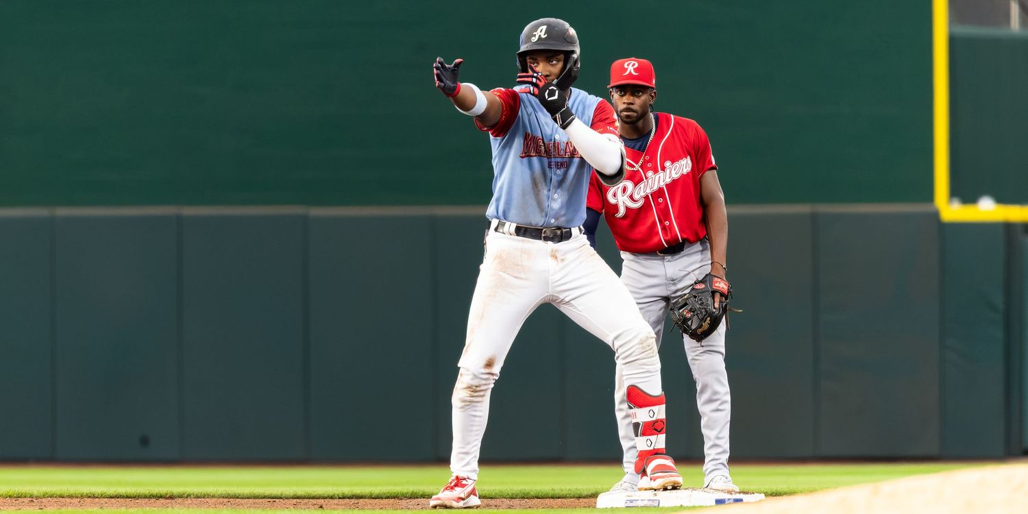 Two baseball players on field, one pointing after reaching base, wearing red and white uniforms during a game.