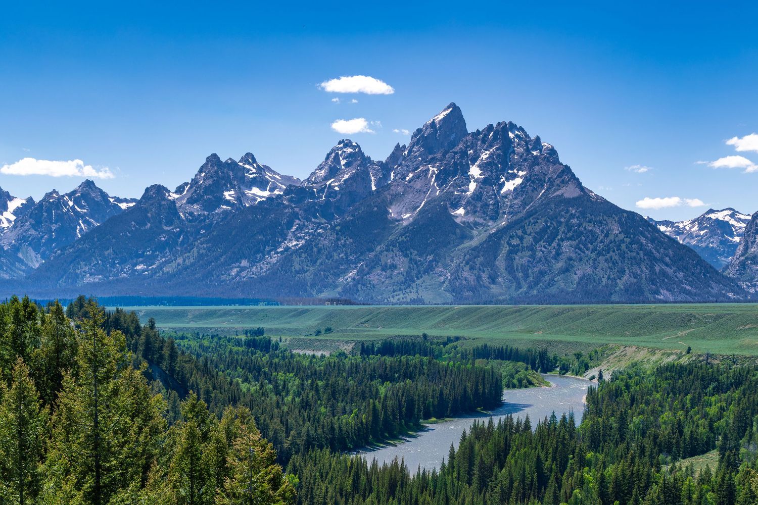 Panoramic view of snow-capped Grand Teton mountain range with evergreen forest and winding Snake River below.