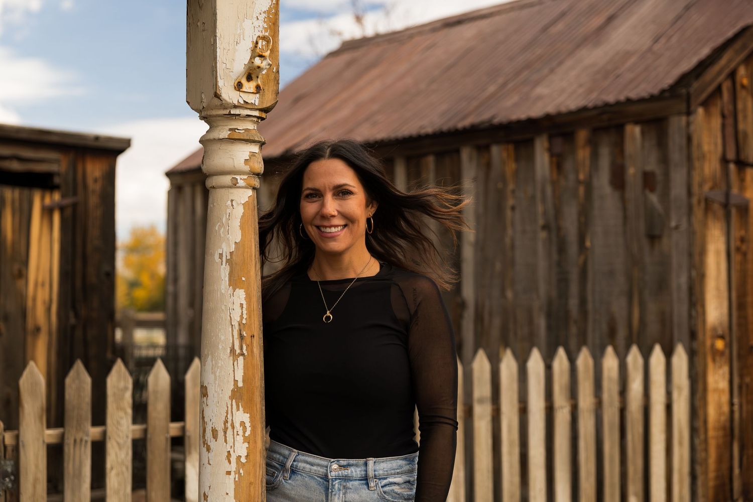 A group of people pose together in front of rustic wooden buildings and fencing during golden hour lighting.