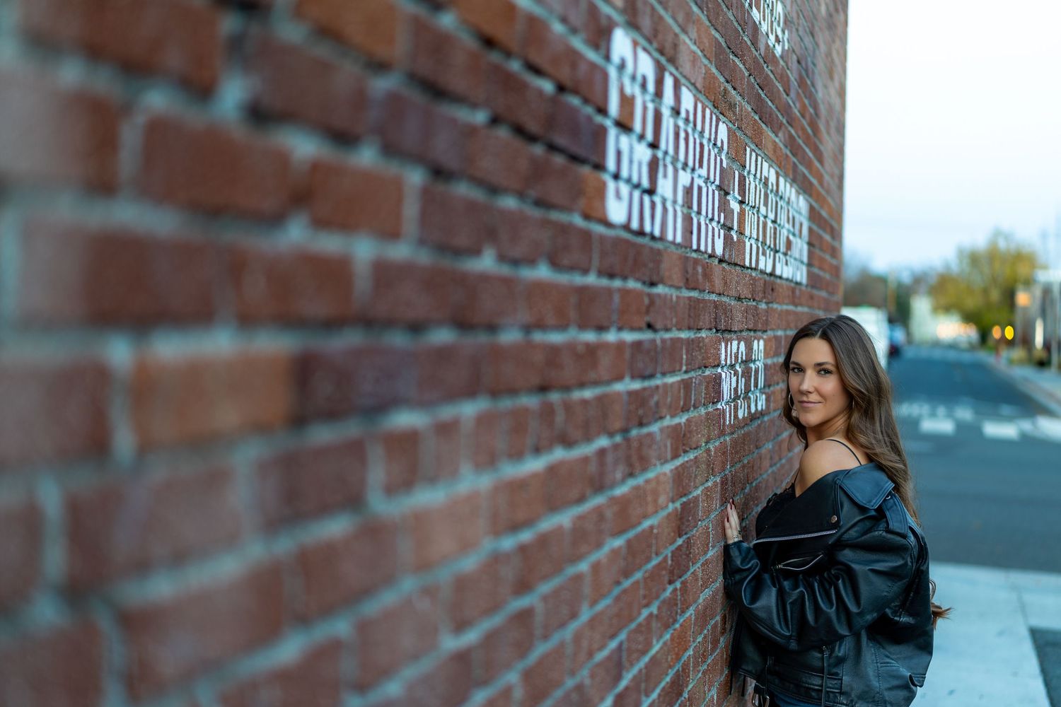 A person in a black leather jacket poses against a brick wall on an urban street corner.