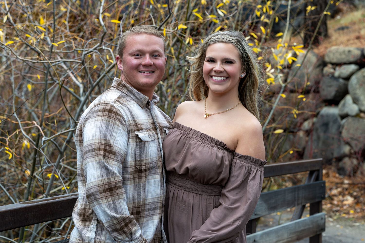 A couple in brown and plaid clothing poses together outdoors among autumn foliage for engagement photos.