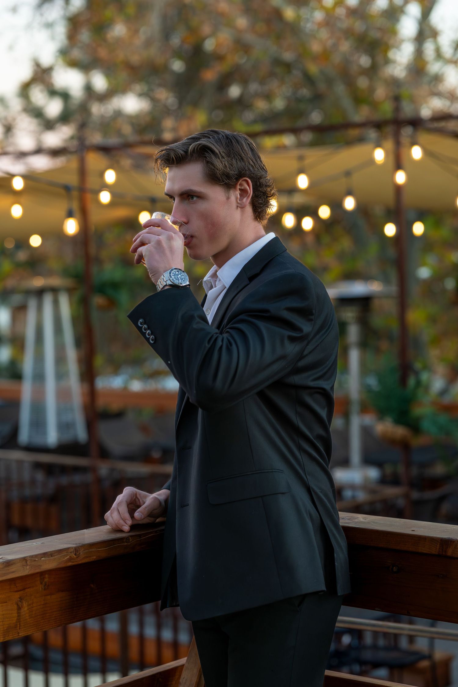 Young man in dark suit sipping a drink on an outdoor patio with string lights at dusk.