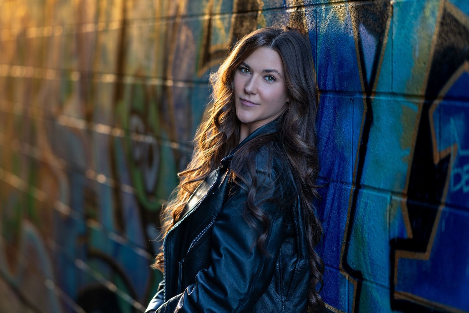 Young woman with long wavy brown hair posing in black leather jacket against colorful graffiti wall.