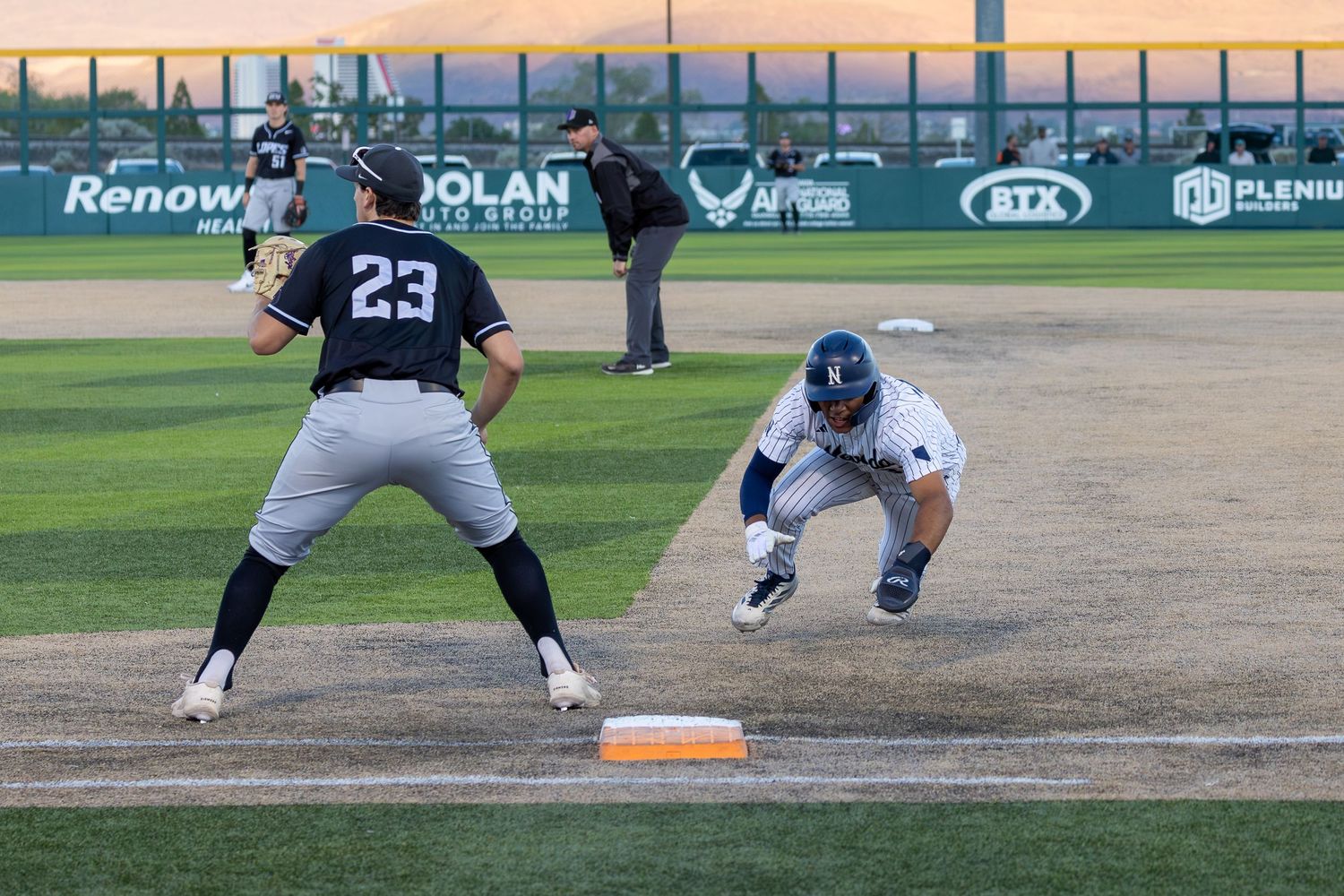 Baseball player #23 dives toward first base as fielder prepares to catch the ball during a competitive game.