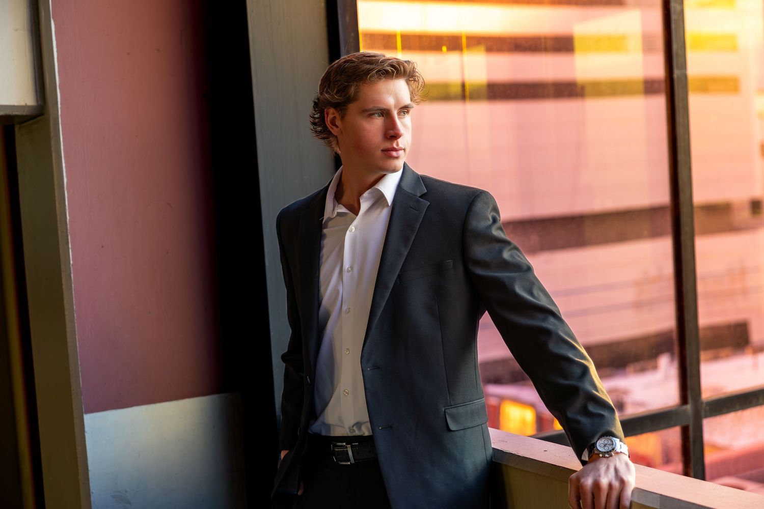 A person in a business suit stands near large windows in an office building during golden hour lighting.