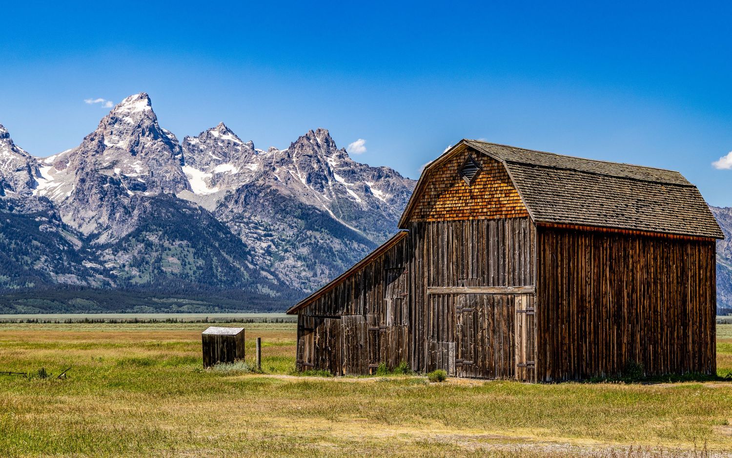 Historic wooden barn stands in field with Grand Teton mountains rising dramatically behind.