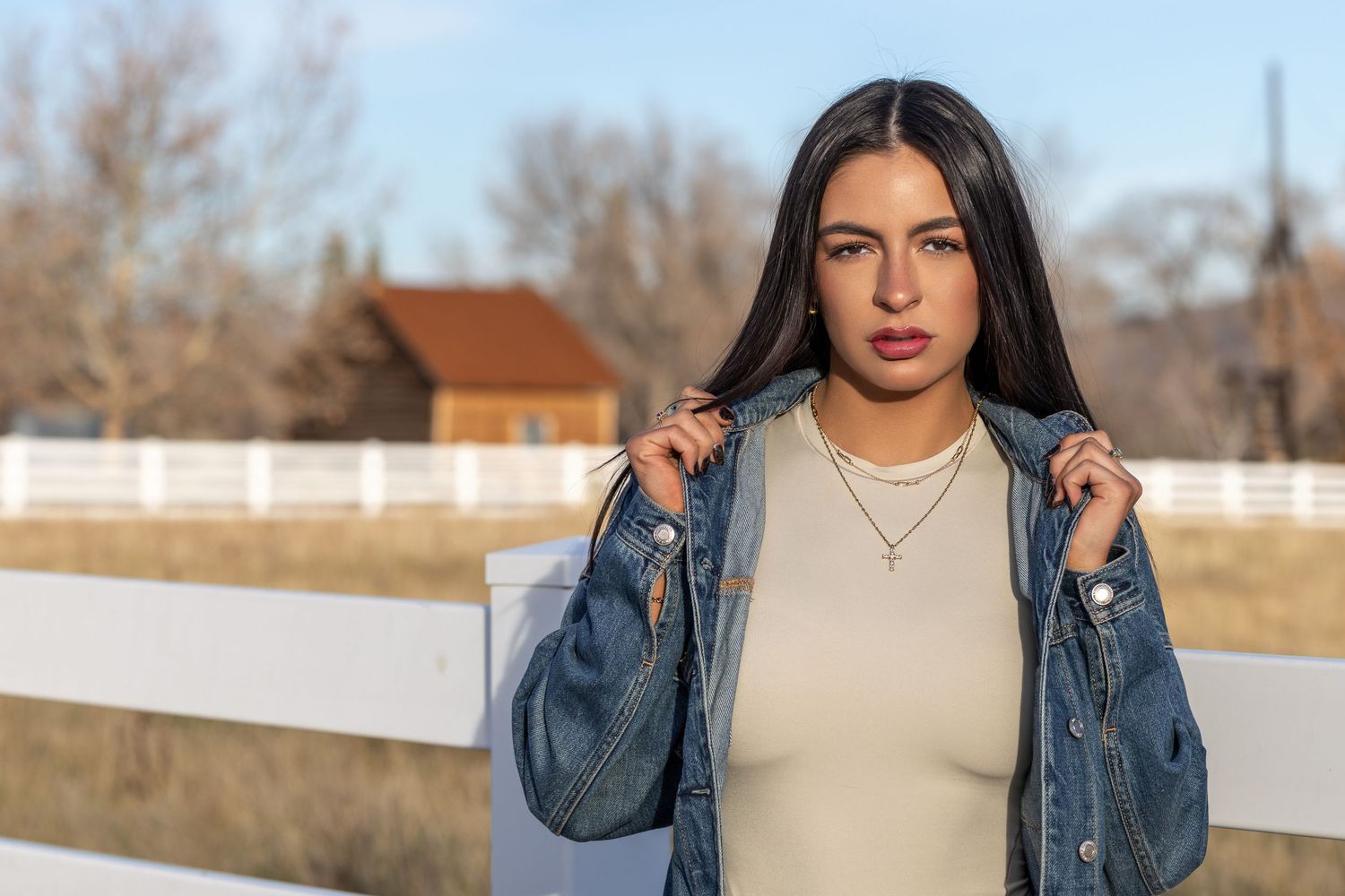 Person in denim jacket and beige top poses against white fence in outdoor winter setting.