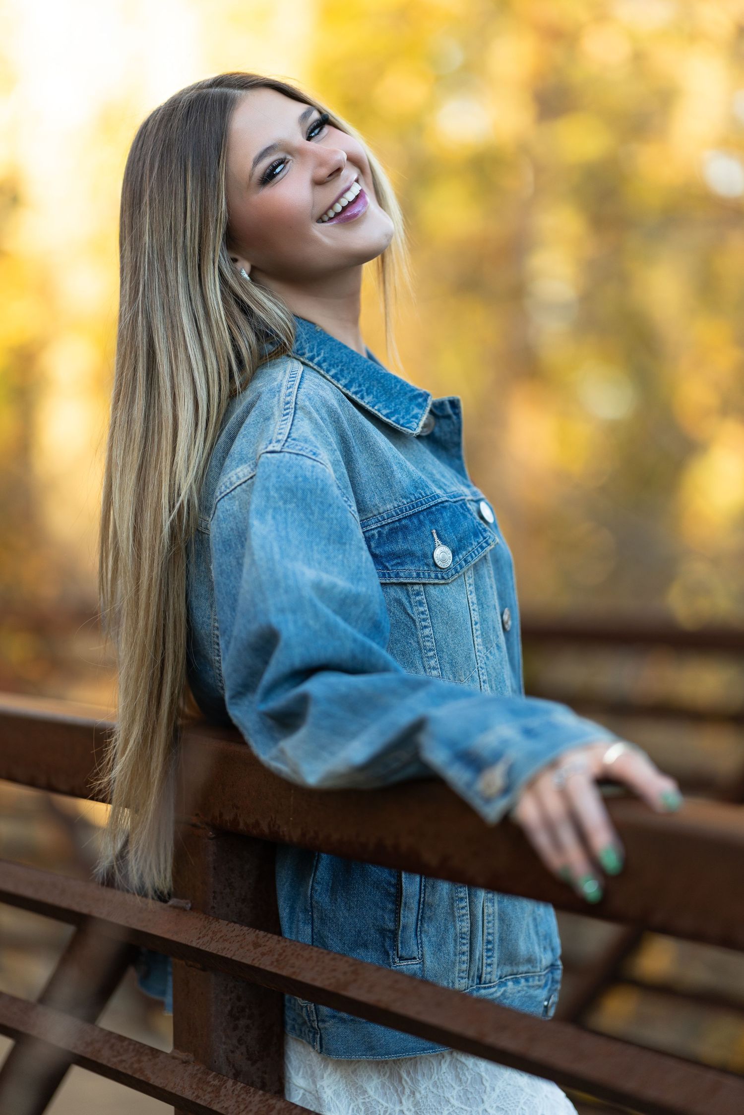 Smiling young woman with long blonde hair wearing an oversized denim jacket, leaning on a rustic fence with autumn foliage.
