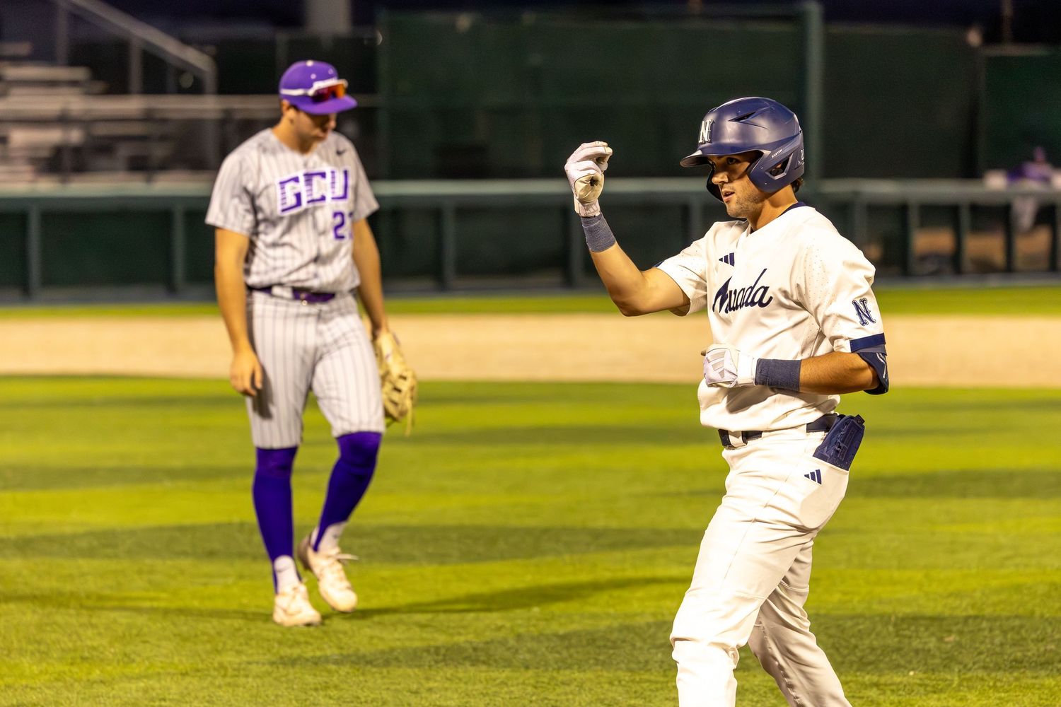 Baseball player celebrates on base while opponent in purple GCU uniform stands nearby during night game.