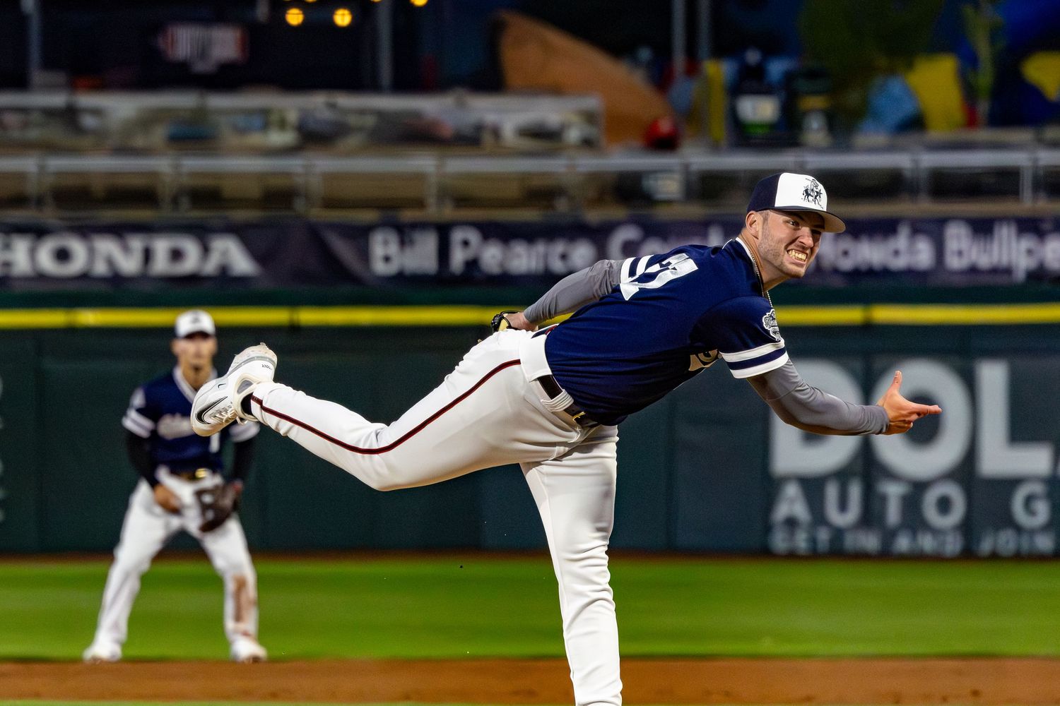Baseball pitcher in navy uniform delivering a pitch at night game with teammate visible in background.