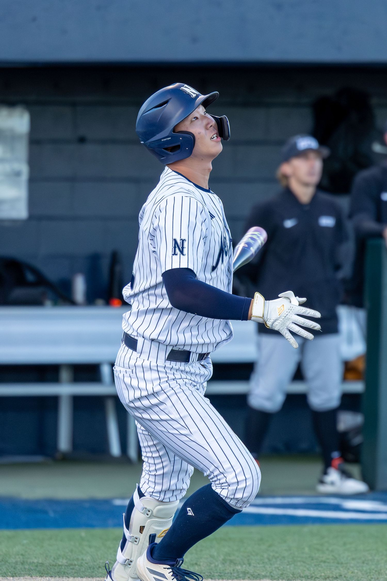 Baseball player in pinstripe uniform looks skyward after hitting, with dugout visible in background.