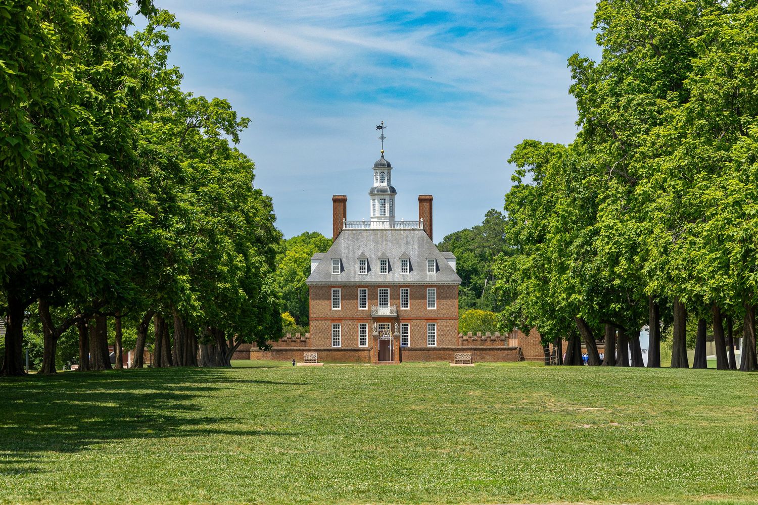 Historic brick colonial Governor's Palace in Williamsburg, Virginia, framed by lush green trees under a bright blue sky.