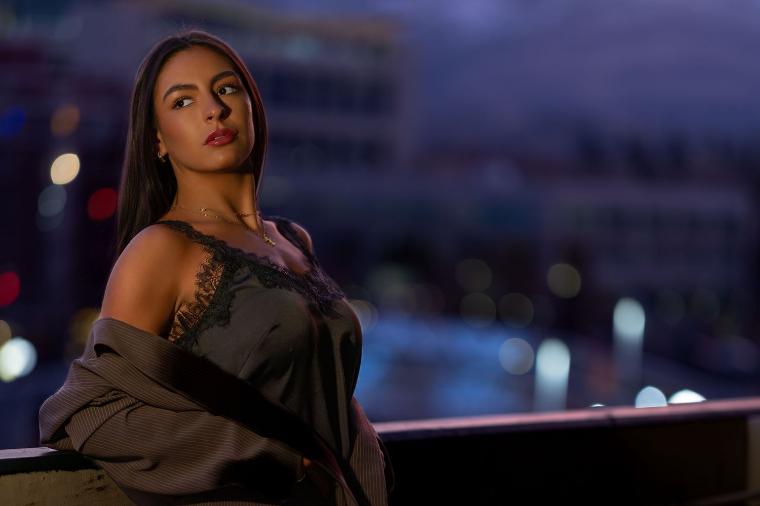 A young woman in a black lace top and gray jacket poses on a rooftop at night with city lights bokeh background.