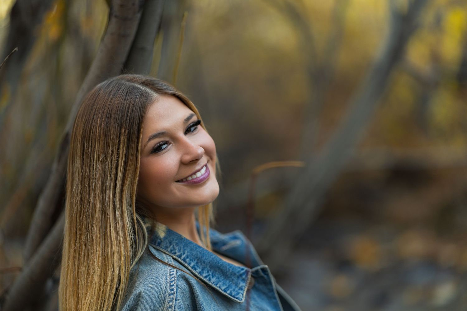 Young woman with long hair smiling in autumn forest, wearing denim jacket, outdoor portrait photography.
