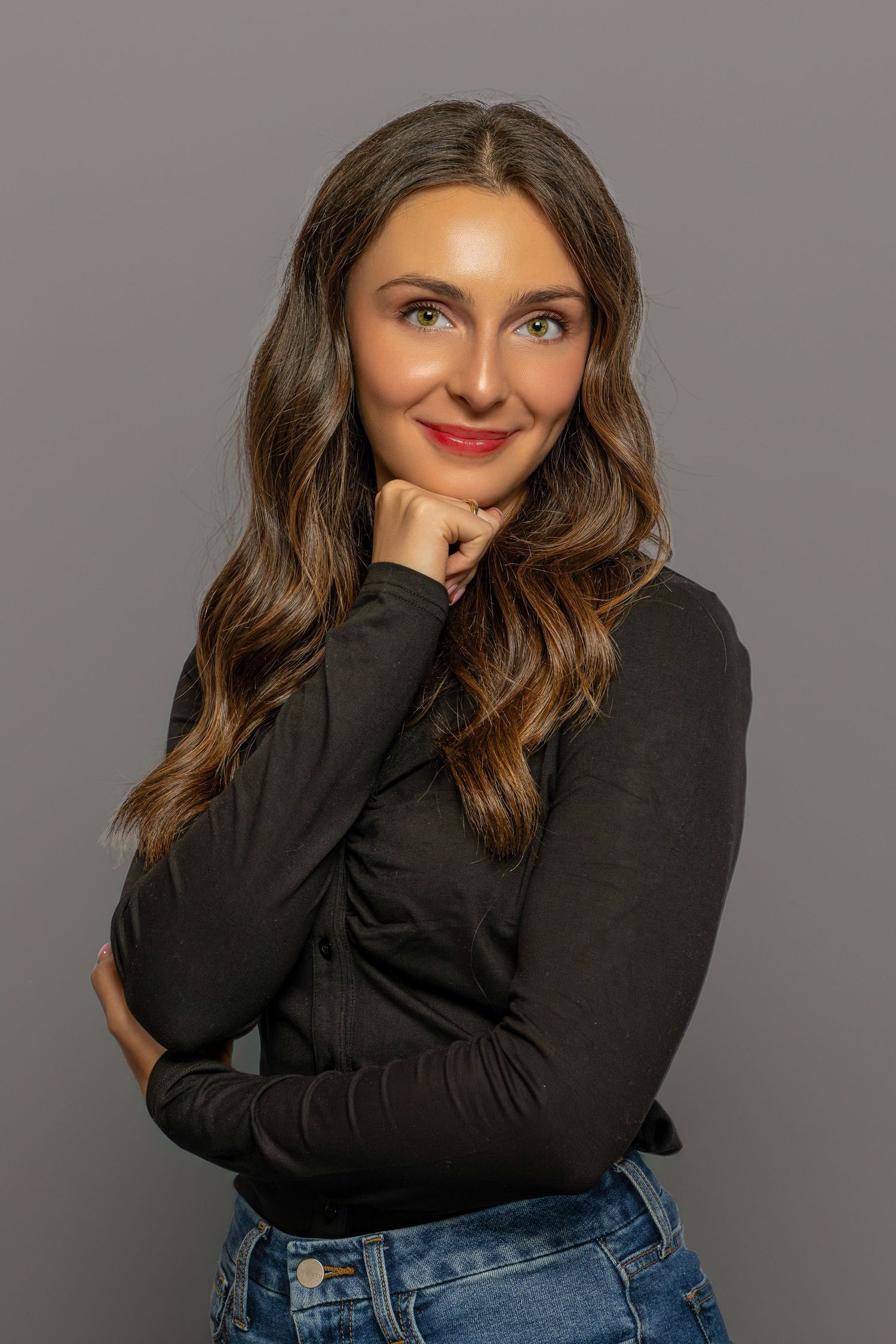 Young woman with long wavy brown hair in black turtleneck and jeans, smiling confidently with hand on chin.