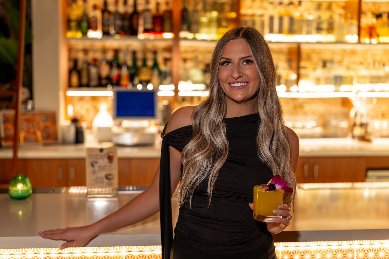 A bartender in a black dress stands smiling at an illuminated bar counter with bottles displayed on shelves behind.
