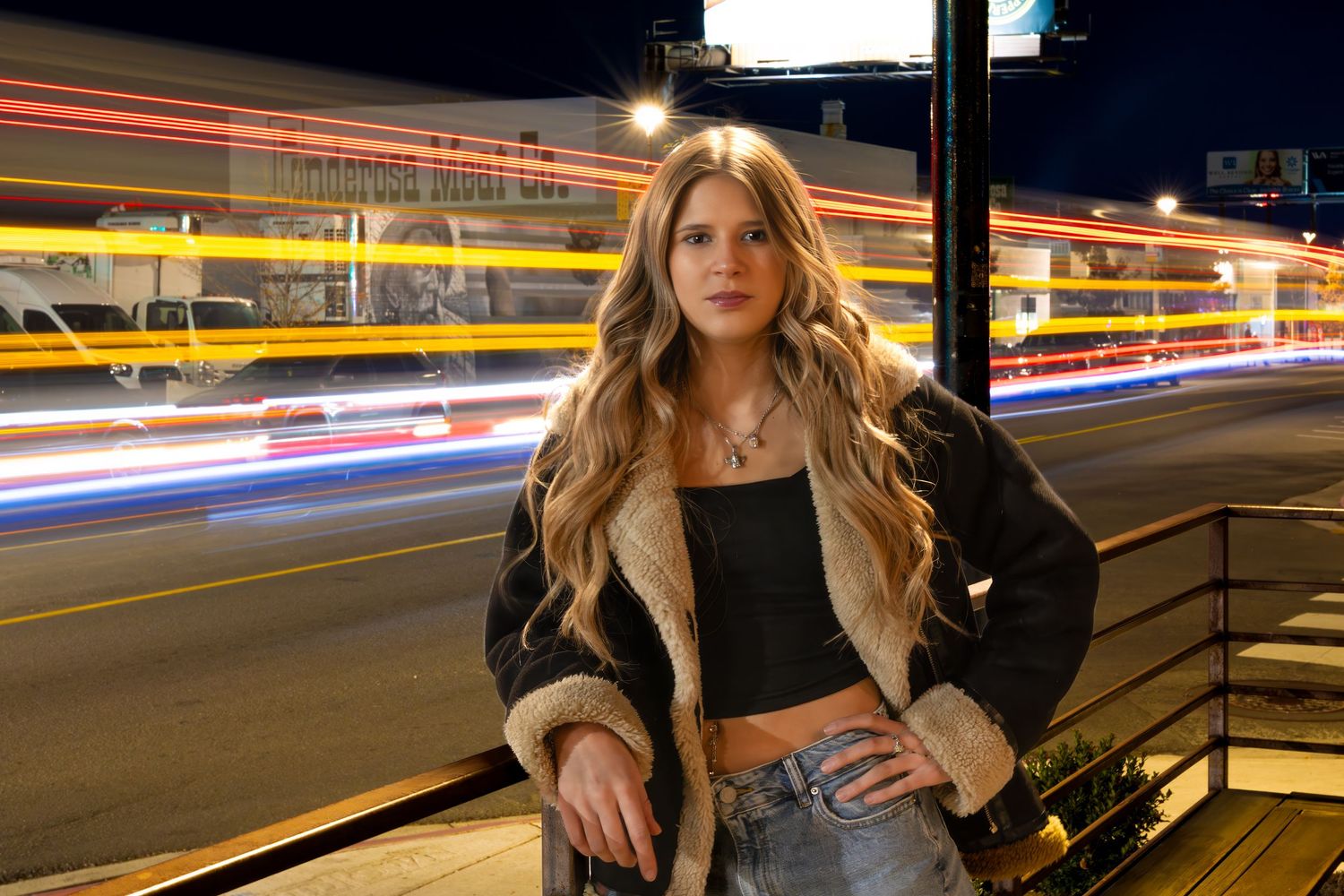 Person with long blonde hair poses against railing at night with streaks of light from passing cars in background.