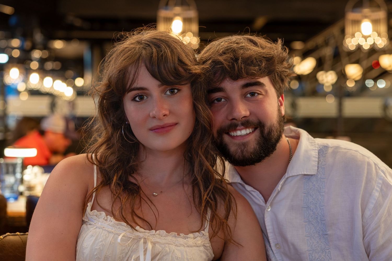 A smiling young couple poses together at a warmly lit restaurant, woman in white dress, man in white shirt.
