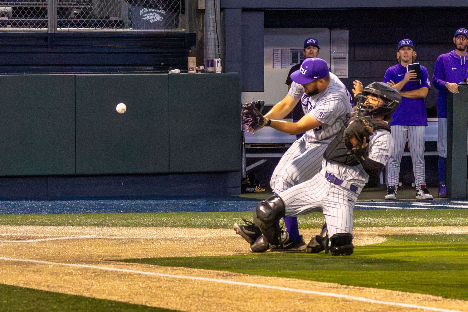 Baseball batter swings at pitch with catcher crouching behind home plate during a night game at a collegiate baseball stadium.