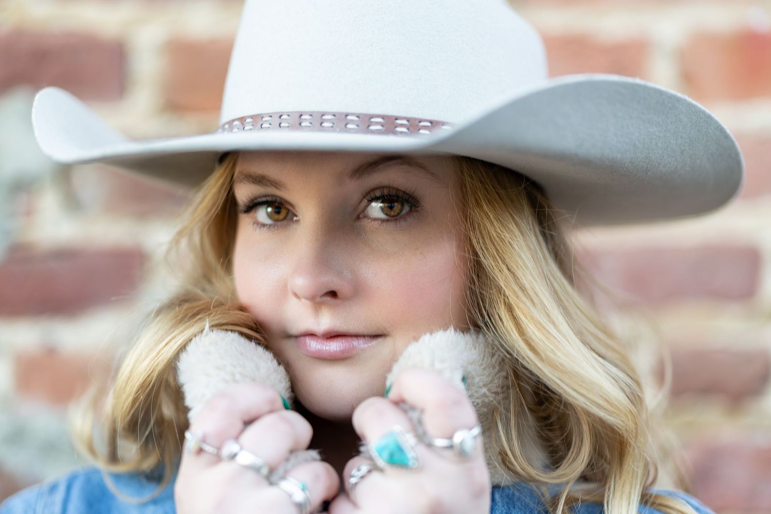 A person wearing a light gray cowboy hat and blue denim jacket holds up soft pink mittens against a brick wall background.