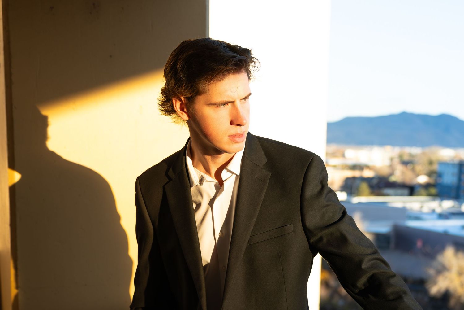 A person in a dark suit jacket stands in dramatic lighting near a window with mountains visible in the background.