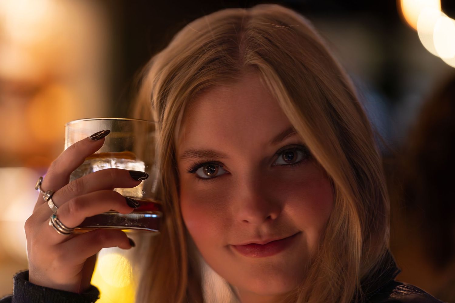 Young blonde woman holding a glass of whiskey, smiling softly in a warmly lit bar setting.