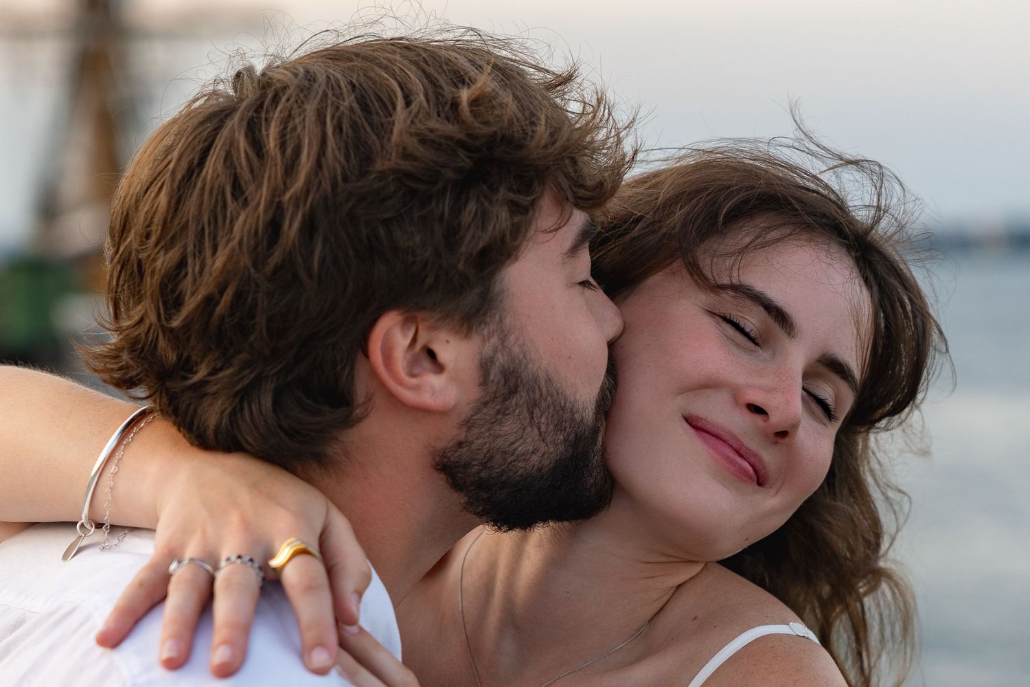 Young couple sharing a tender moment, man kissing woman on cheek near waterfront at sunset.