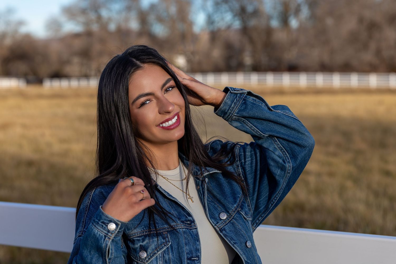 A woman in a denim jacket smiles and poses outdoors during golden hour in a grassy field.