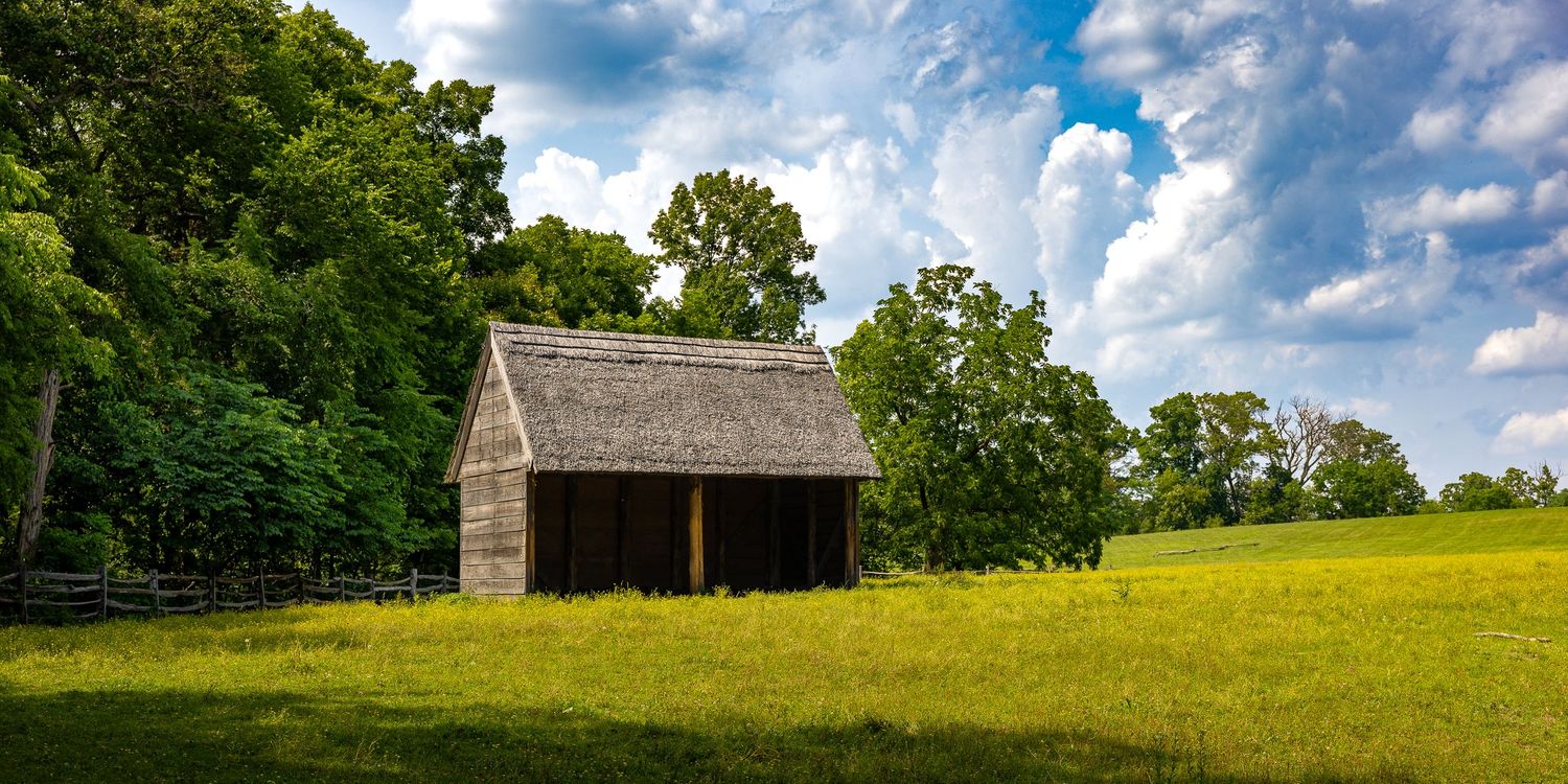 A rustic wooden barn sits in a lush green meadow surrounded by tall trees under a bright blue sky with fluffy clouds.