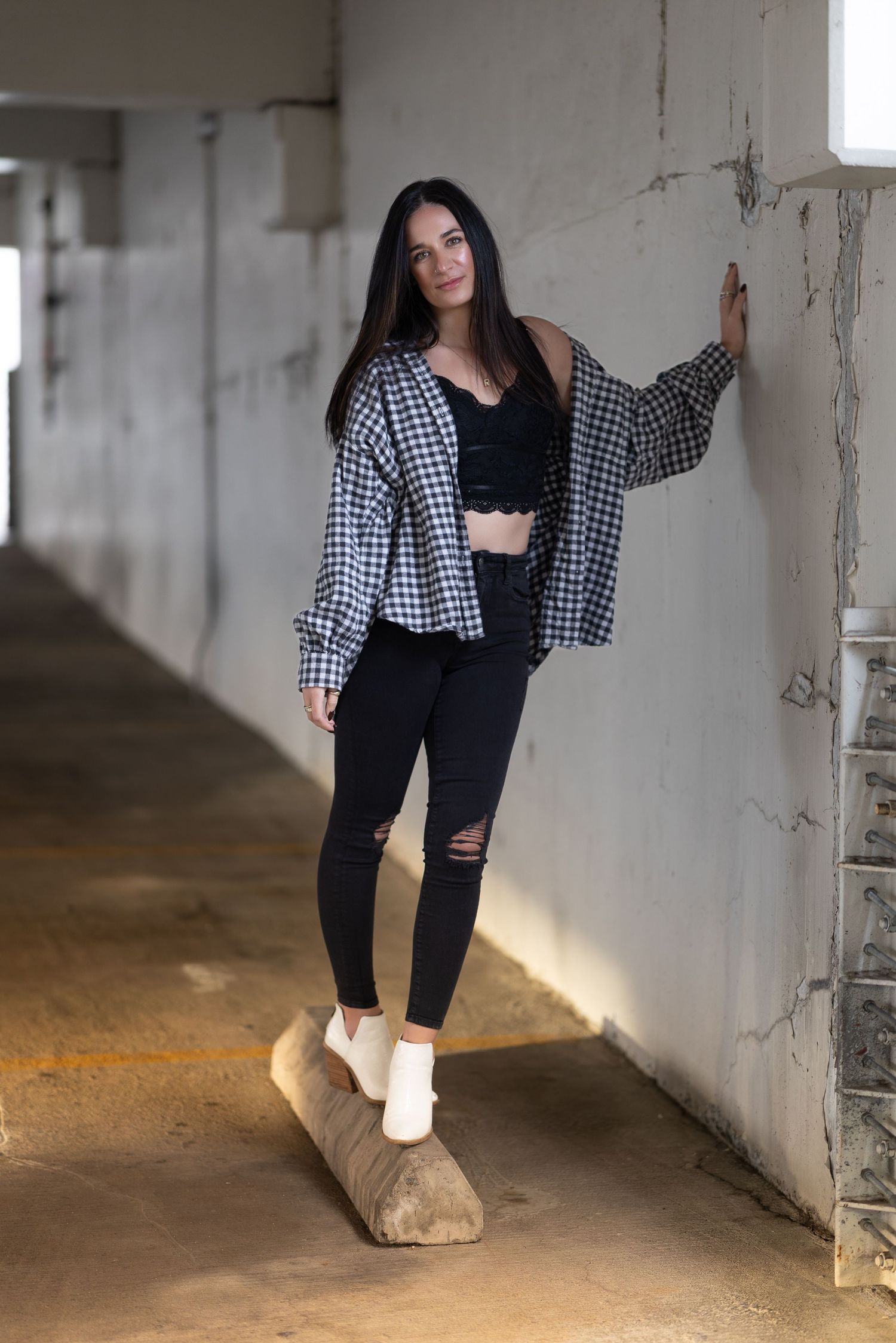 A person in a black and white checkered shirt and dark pants poses in an industrial concrete hallway.