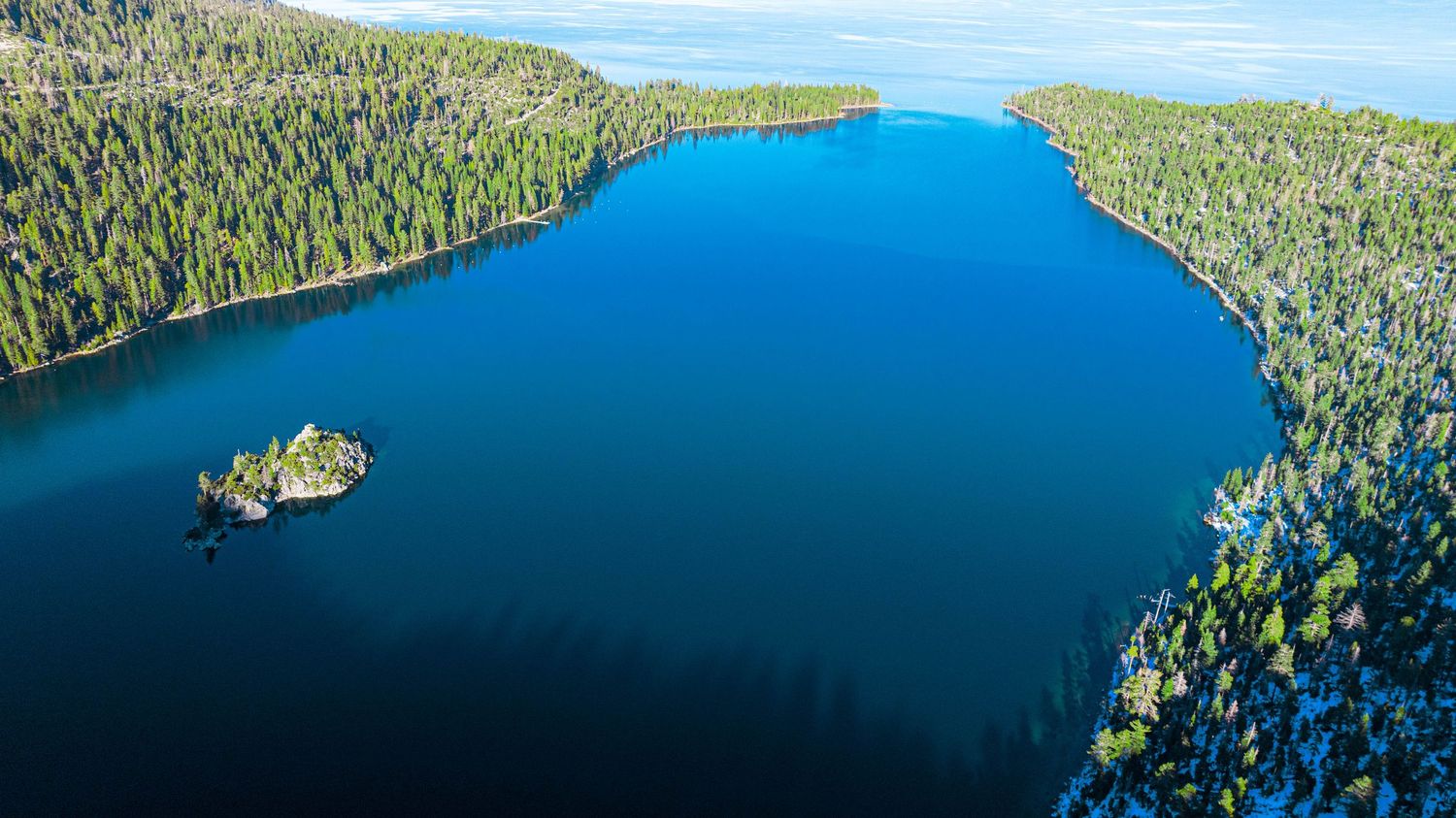 Aerial panoramic view of a pristine mountain lake surrounded by dense evergreen forest and rocky shoreline.