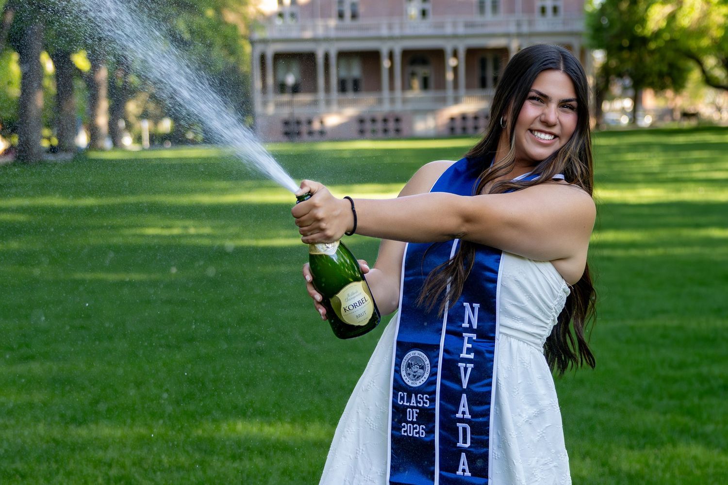 University of Nevada graduate celebrates in blue sash, spraying champagne on green campus lawn.