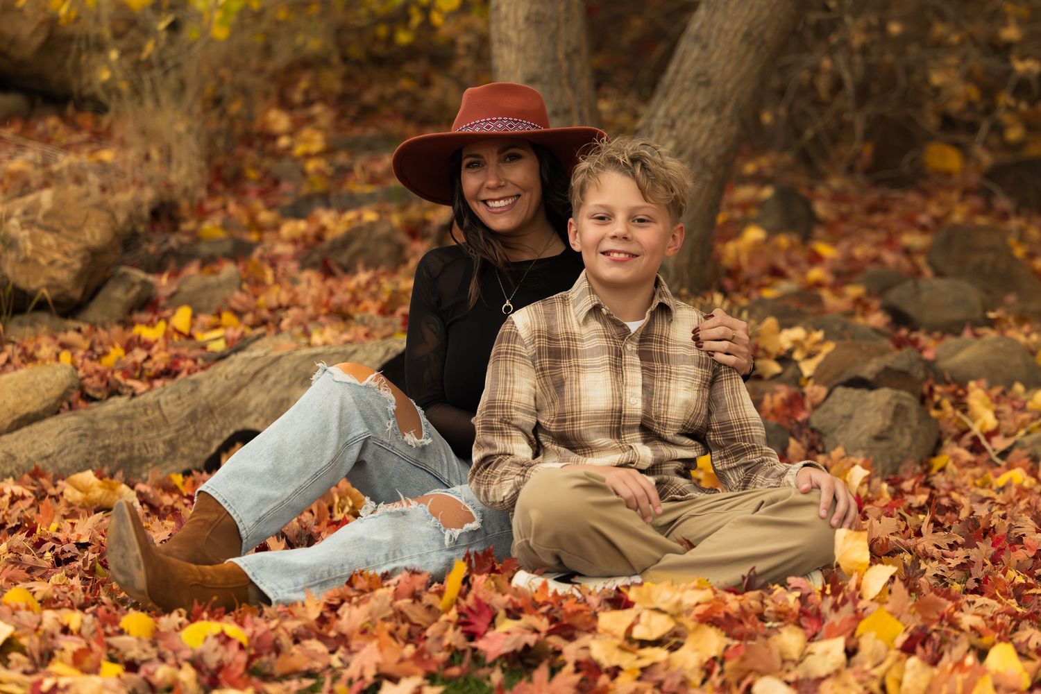 Two people sit together on fallen autumn leaves in a park wearing casual clothing and enjoying the fall season.