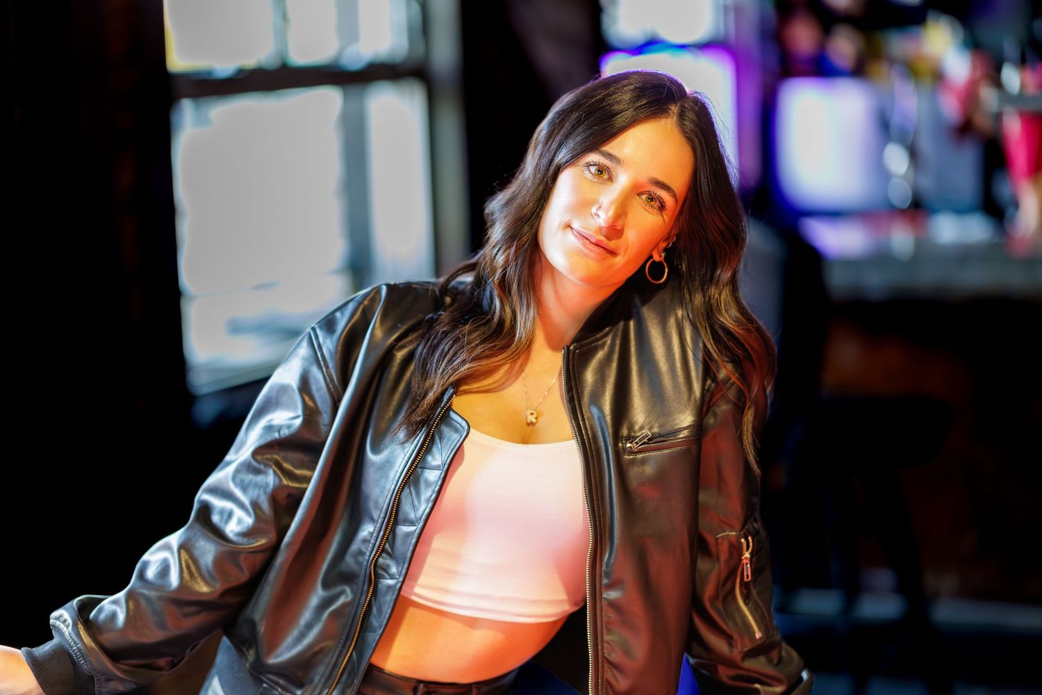 Woman in leather jacket poses against colorful neon lights in urban nighttime setting.