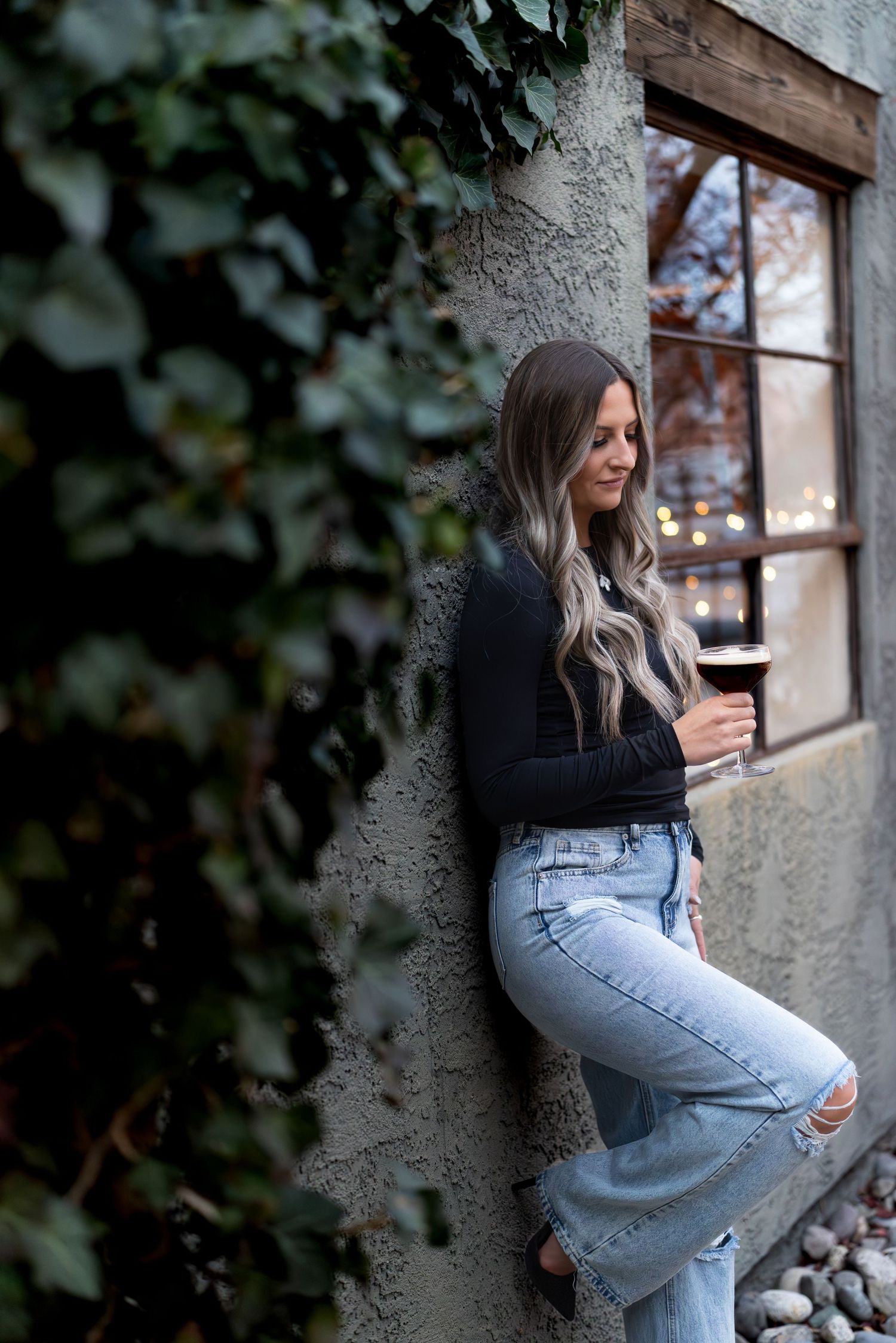 A person in a black sweater and light jeans poses against a textured wall with windows and foliage in the background.