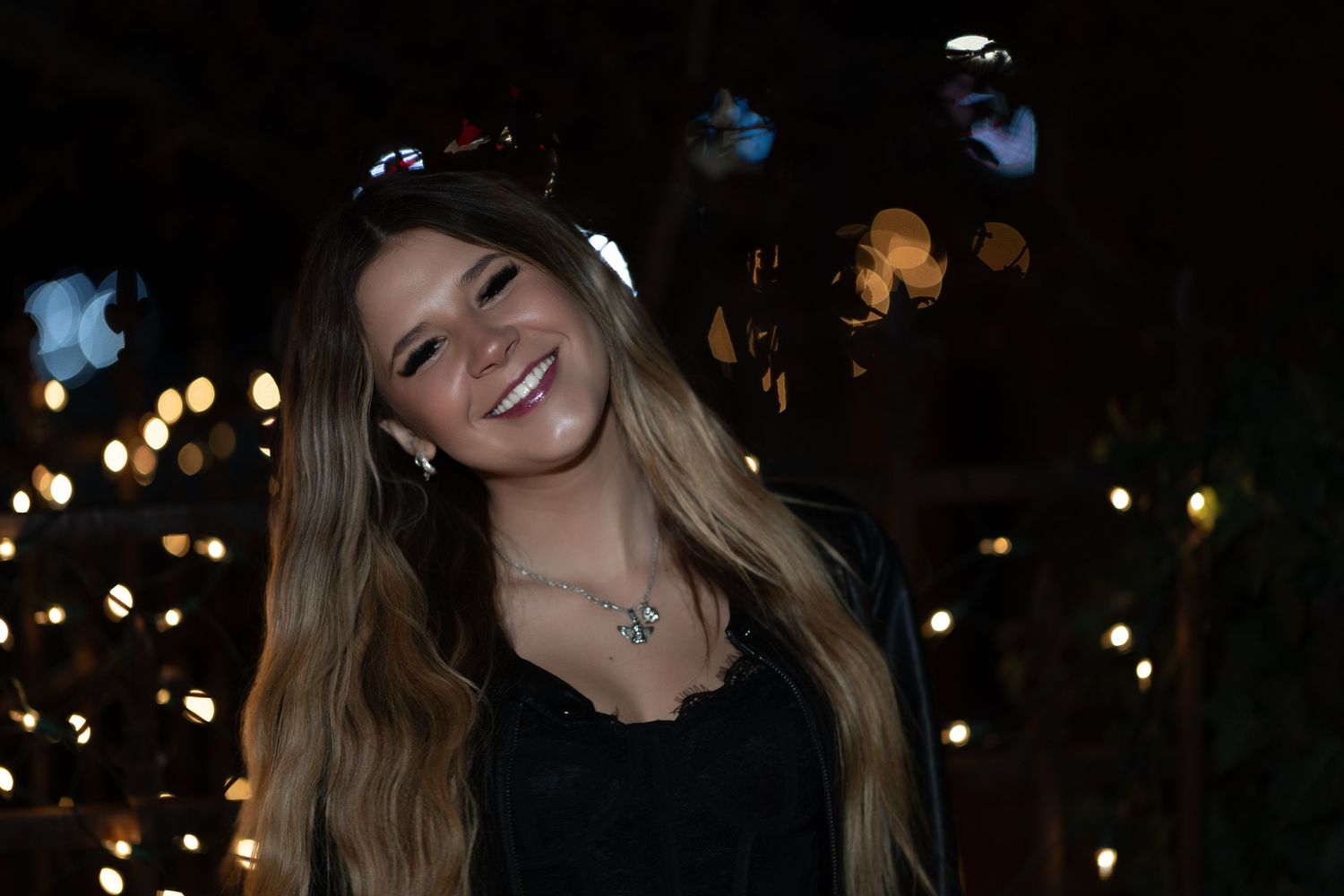 Smiling young woman with long hair and sunglasses on head, wearing black top and necklace, bokeh lights background.